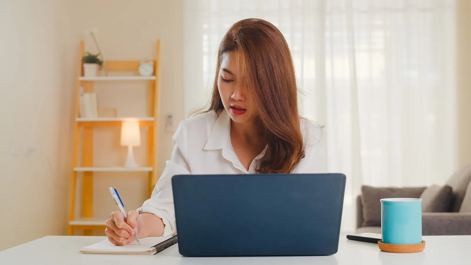 A person taking notes while working on a laptop at home